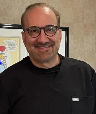 Male orthodontist wearing glasses and black shirt smiling in office setting.