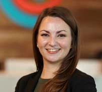 Professional headshot of smiling woman with long dark hair in black blazer against blurred office background.
